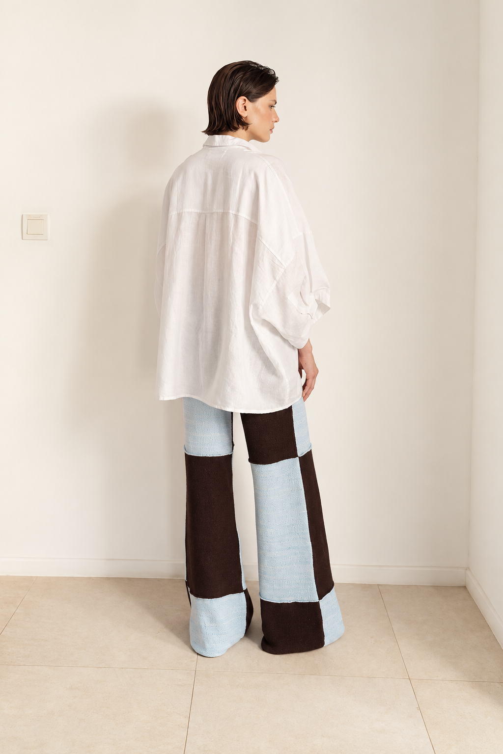Woman wearing a checkered bottoms with ice blue and coffee brown large checker pattern and raw seams. On top she is wearing a white linen shirt with 3/4 sleeves.