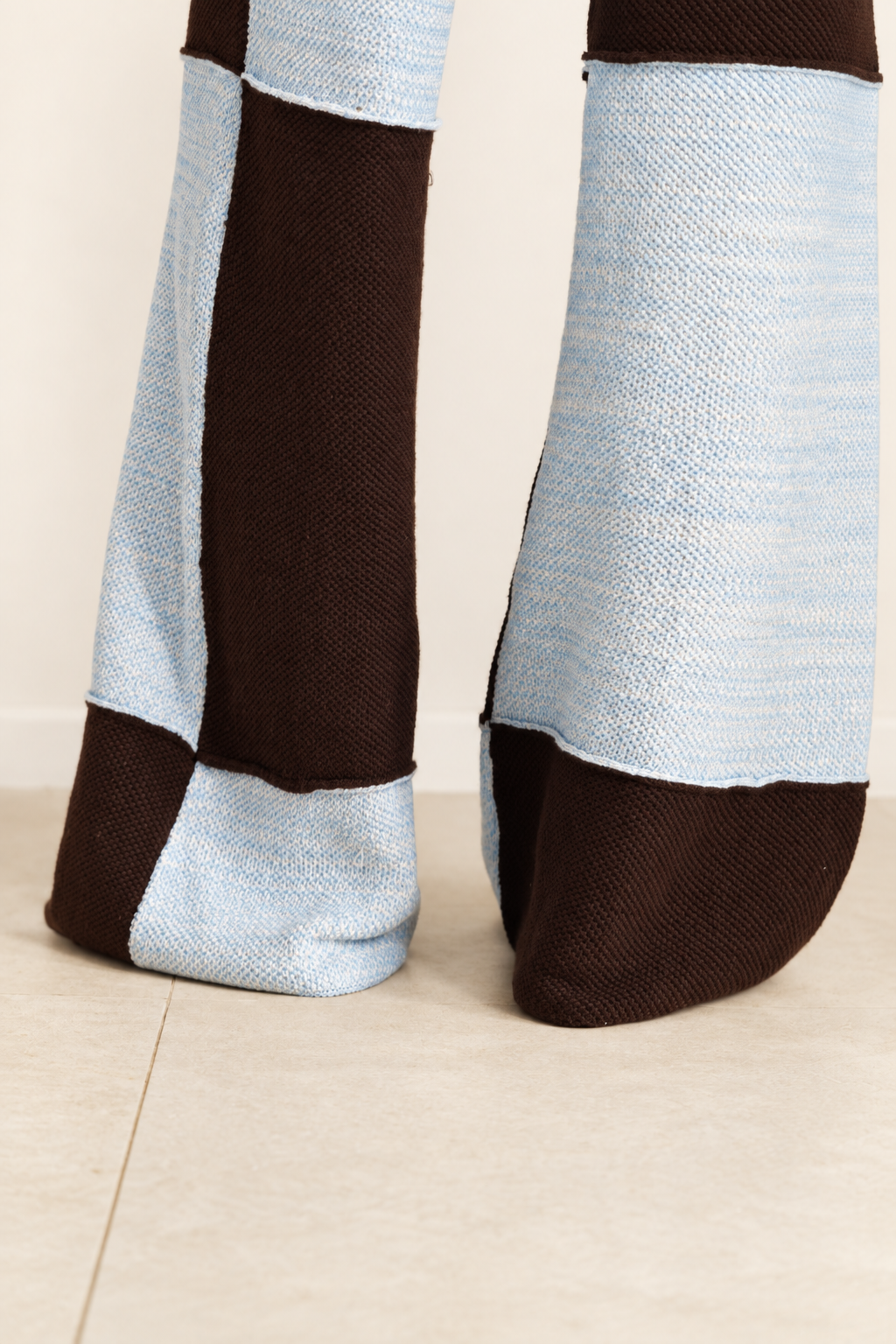 Close-up of a person's feet wearing knit socks with a color-blocked pattern of light blue and dark brown, standing on a light beige tiled floor.