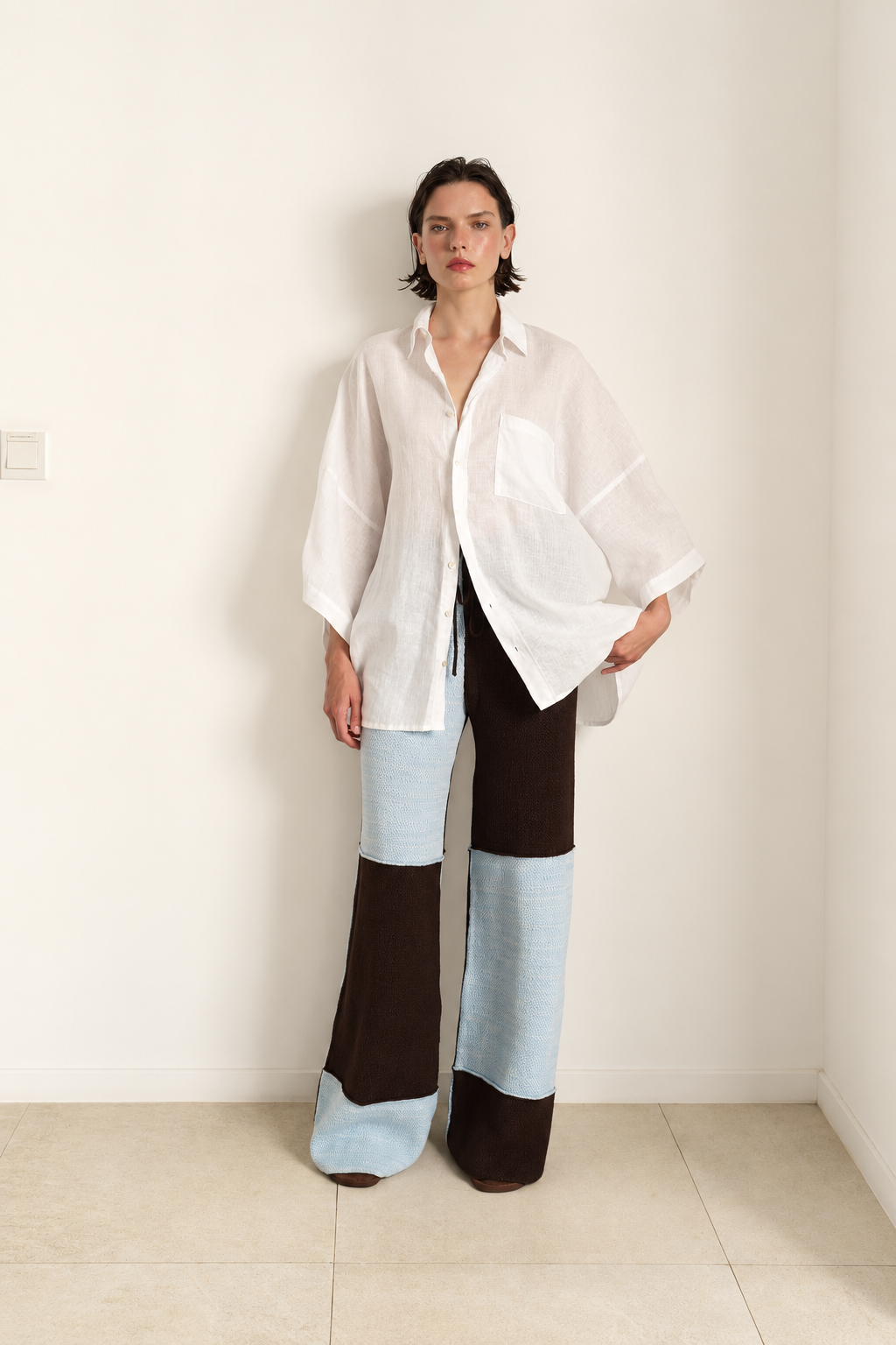 Woman standing against wall wearing a large checkered pants in ice blue and coffee brown, together with a white linen shirt with 3/4 sleeves and a front pocket on chest.