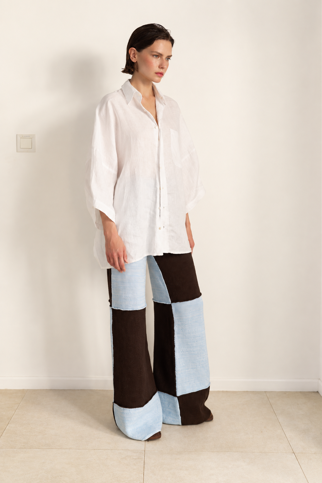 A model stands in a minimalist room wearing a loose white button-up shirt and wide-leg patchwork pants with light blue, dark brown, and beige blocks.