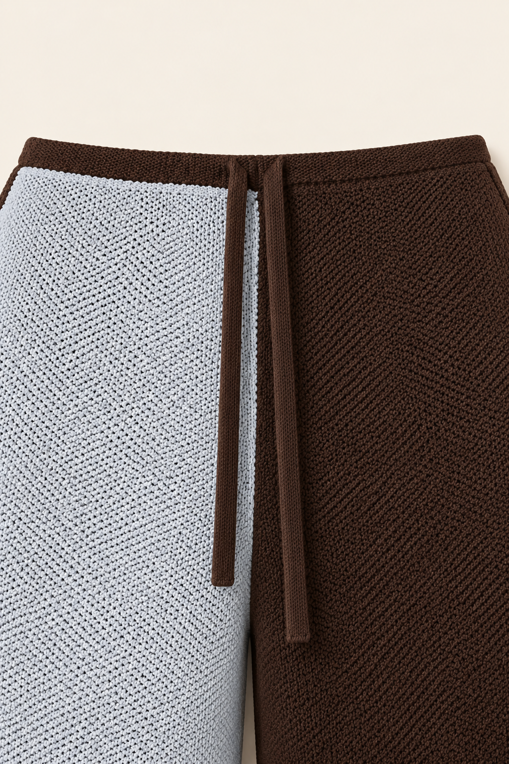 Close-up of a textured knit garment, likely a sweater or pants, split vertically into two colors: a light icy blue on the left and a rich dark brown on the right, with a matching dark brown drawstring at the waist.