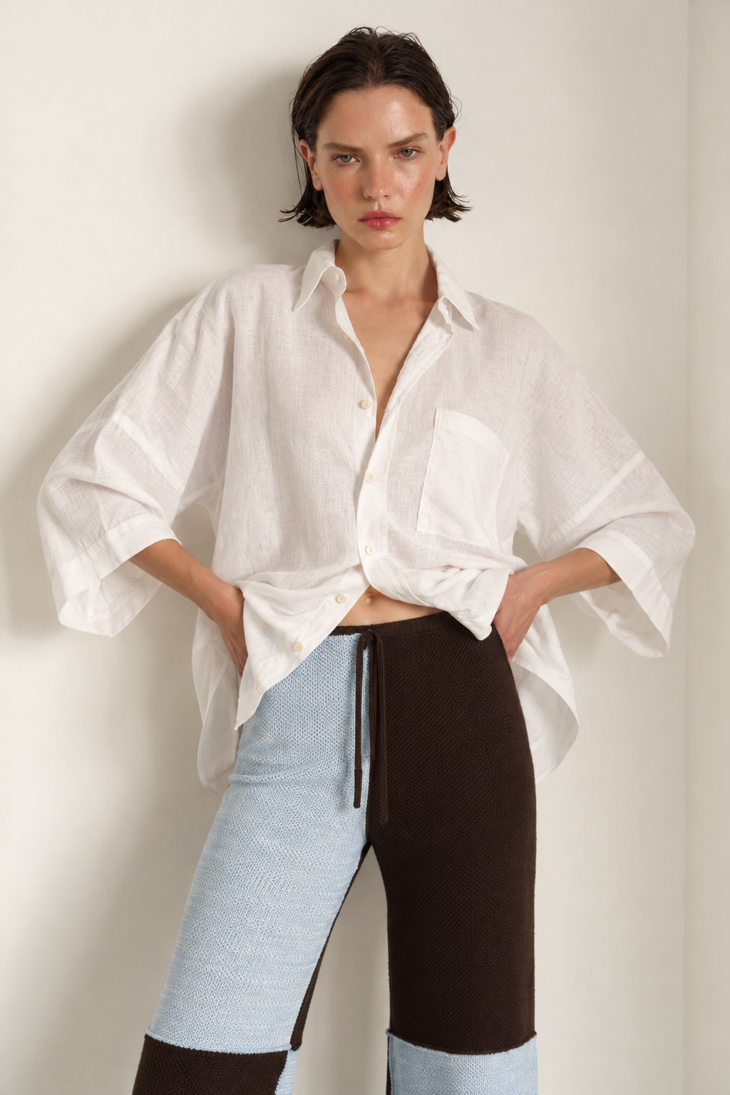 A model wearing a loose white linen button-up shirt and color-blocked knit pants in light blue, dark brown, and light blue, standing against a plain off-white wall.