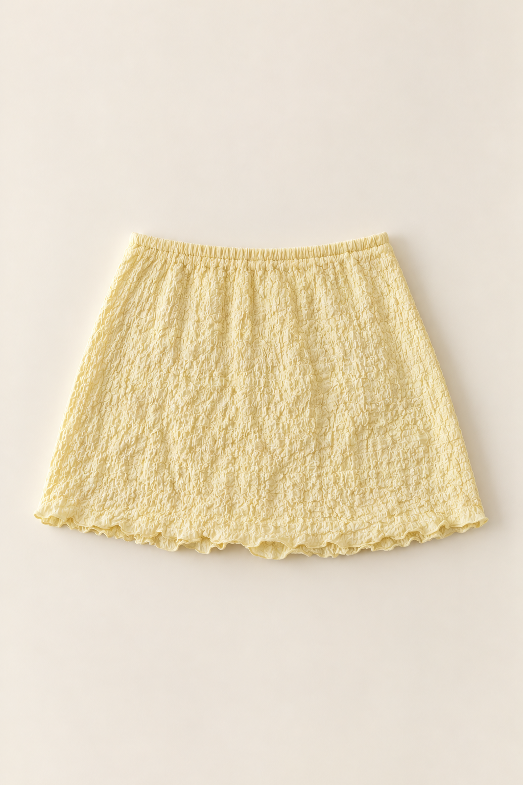 A light yellow textured mini skirt with an elastic waistband and ruffled hem, laid flat against a warm off-white background.
