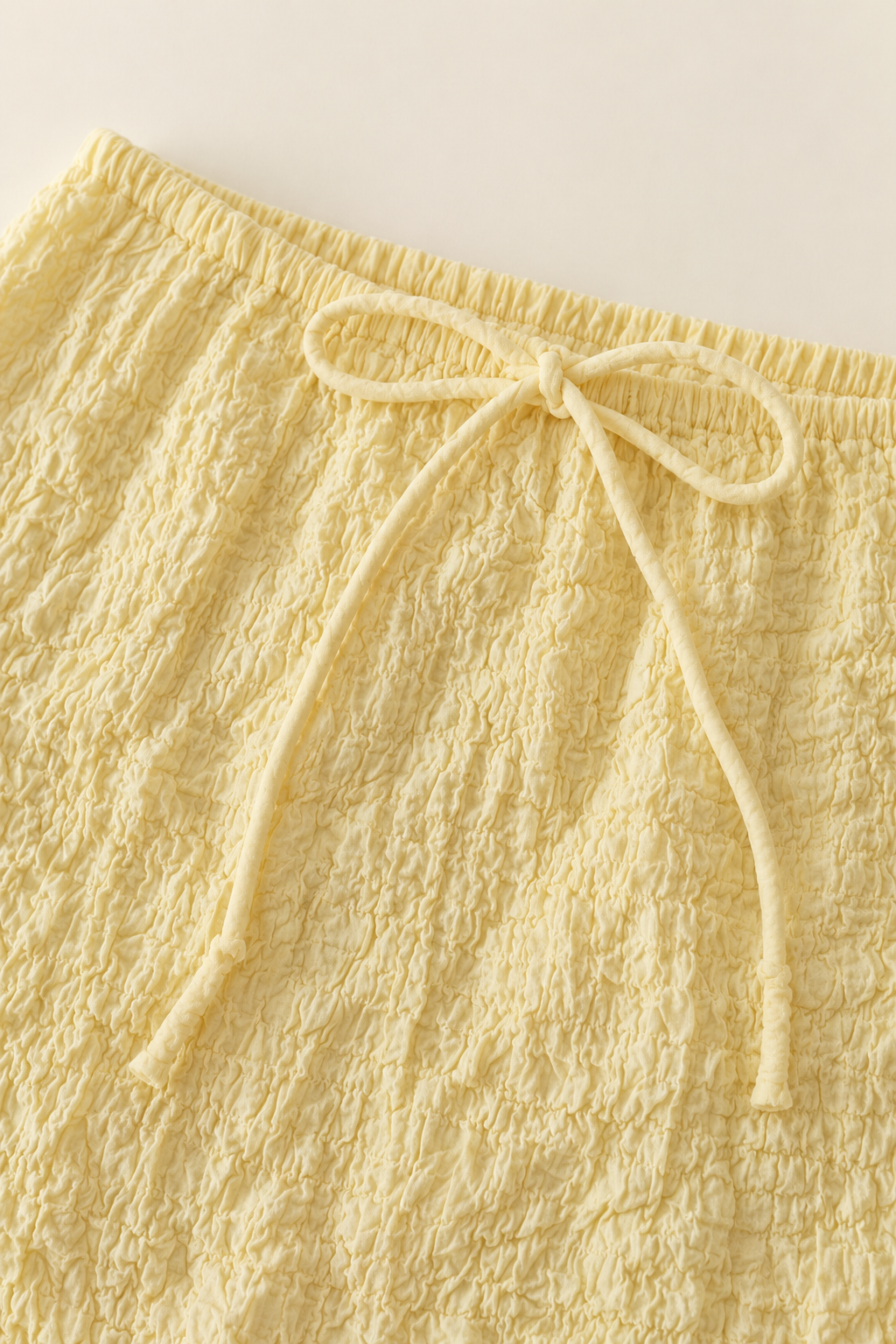 Close-up of pale yellow textured fabric with a matching drawstring tied in a bow at the waistband, photographed against a light neutral background.