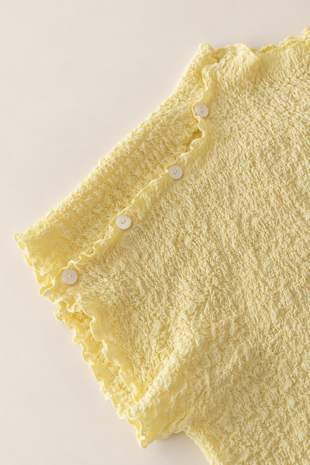 A close-up of a pale yellow textured fabric garment, showing a ruffled edge with small white buttons, laid flat on a light beige background.