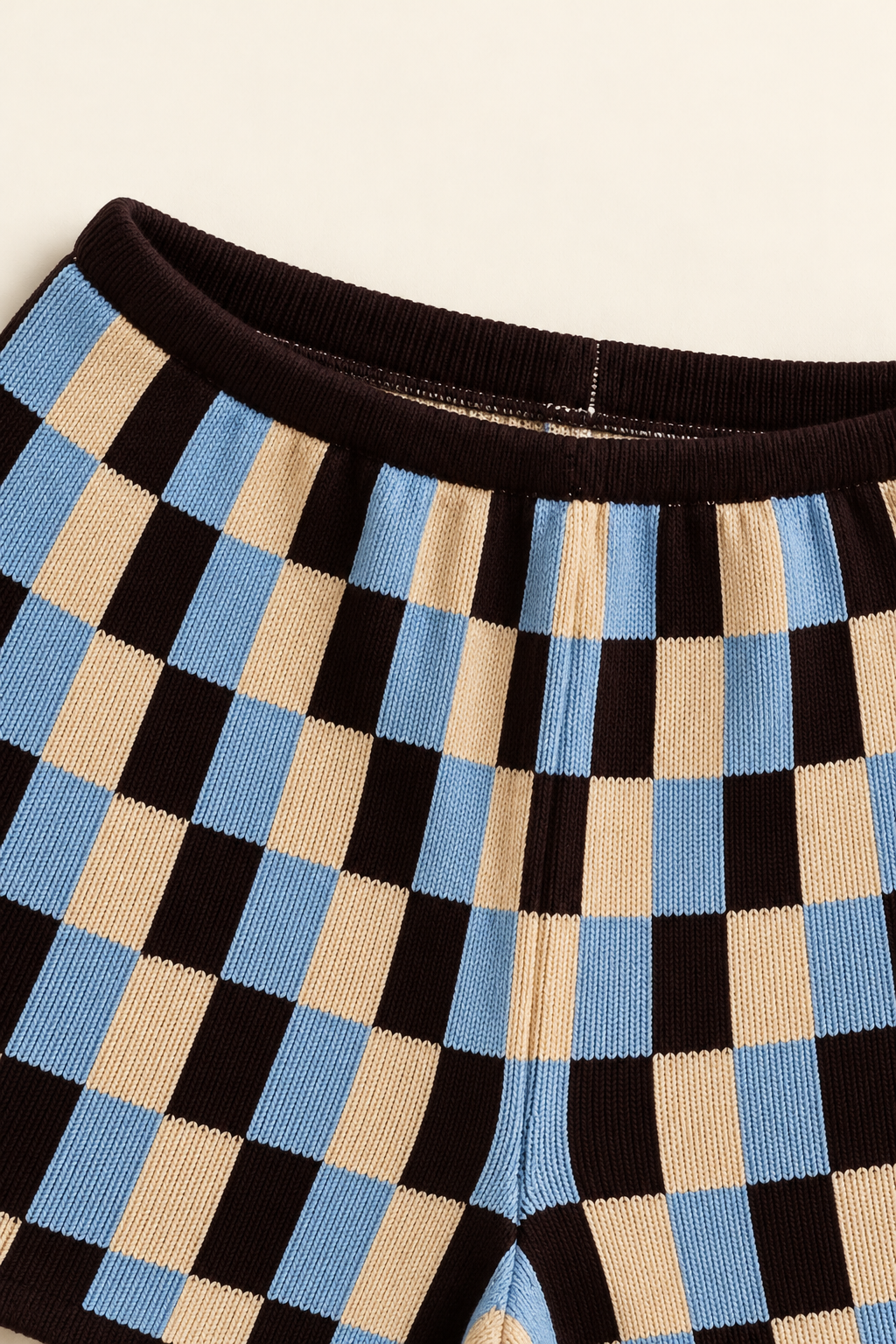 Close-up of a pair of knit pants with a black, beige, and light blue checkerboard pattern and a dark brown ribbed waistband.