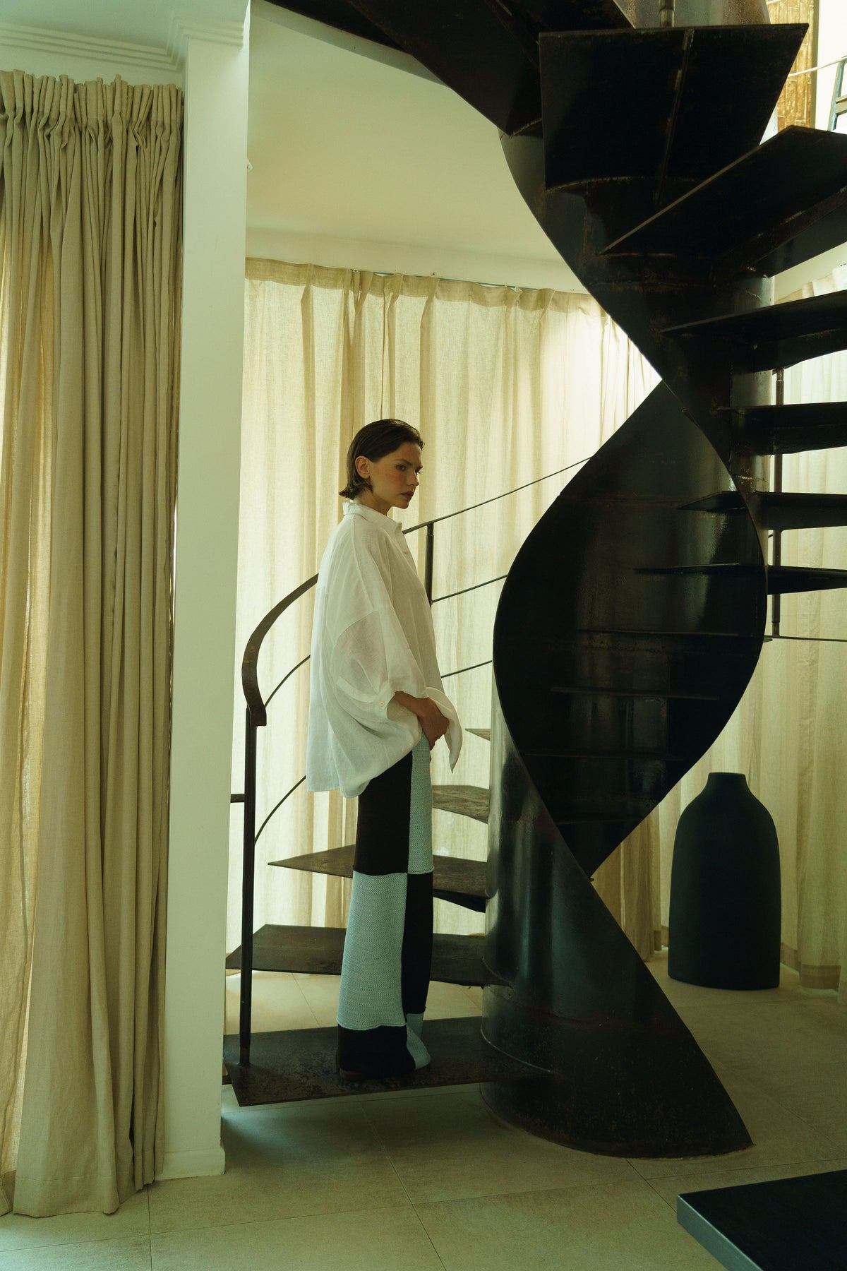 A woman stands on the stairs of a black spiral staircase in a modern interior, wearing a loose white top, black pants, and light blue socks.